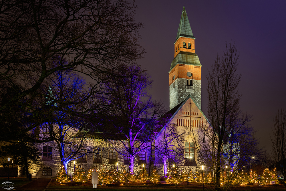 Nachtaufnahme einer Kirche mit Glockenturm, blau-violetter Baumbeleuchtung und Weihnachtslicht vor dem Gebäude.