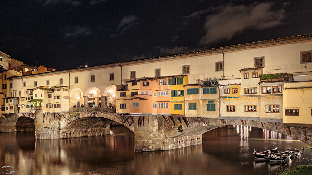 Nachtaufnahme des Ponte Vecchio in Florenz: bunte Häuser am Arno, warm beleuchtet, historische Brücke.