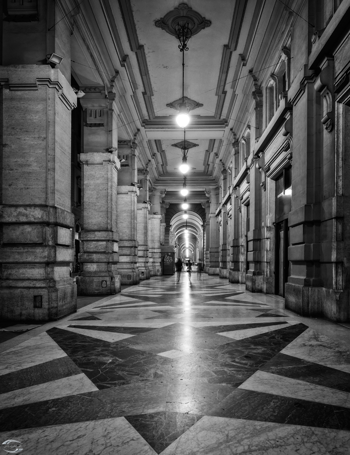 Schwarzweißaufnahme einer historischen Arkadenhalle mit Säulen, Marmorboden im geometrischen Muster und Hängelampen.