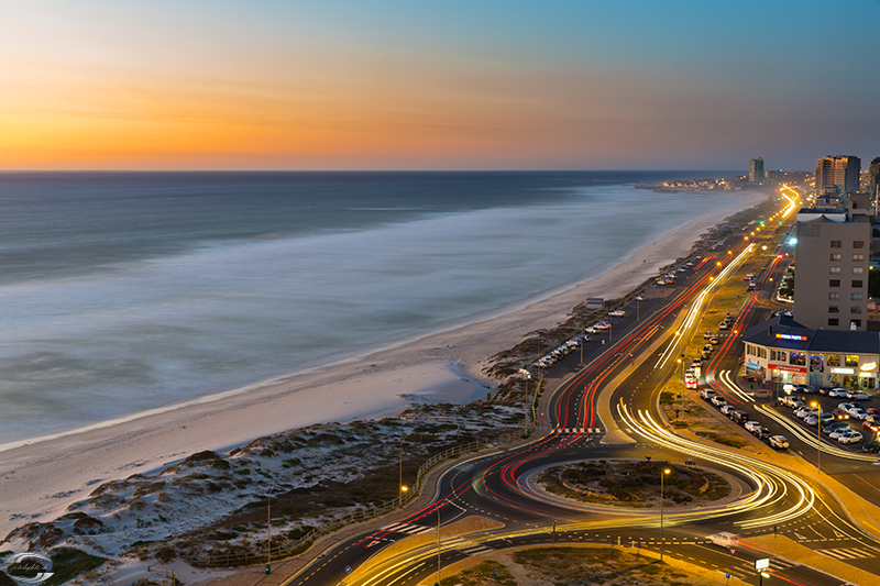 Sonnenuntergang an der Küste: Strand, Meer und beleuchtete kurvige Küstenstraße mit Hochhäusern.