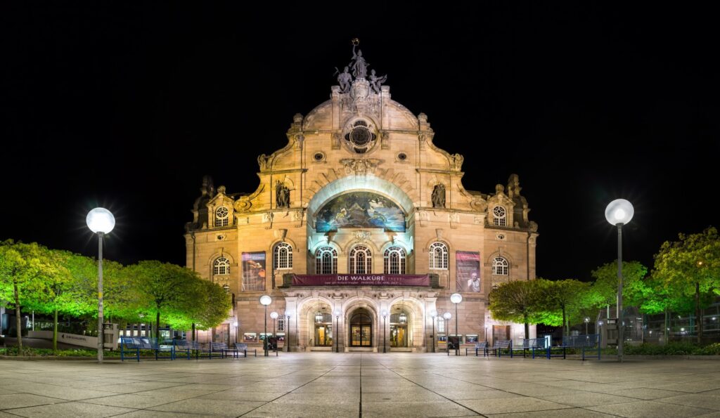 Nachtaufnahme eines historischen Opernhauses mit beleuchteter Fassade, Banner Die Walküre und hellem Stadtplatz.