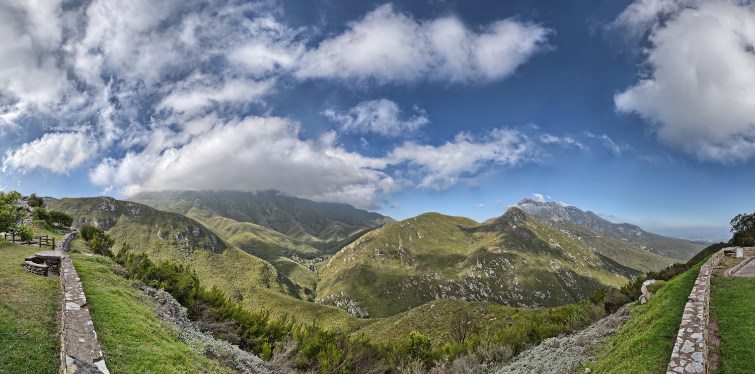 Panorama grüner Berglandschaft mit Steinpfad, grasbewachsenen Hügeln und wolkenverhangenem blauen Himmel