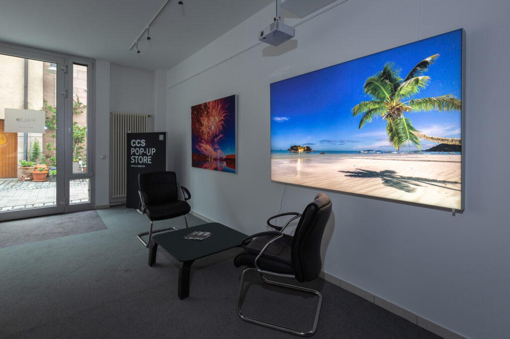 Moderne Pop-up-Store Lounge mit zwei schwarzen Ledersesseln, Strandmotiv-TV und Feuerwerkbild an der Wand, Glas-Eingang.