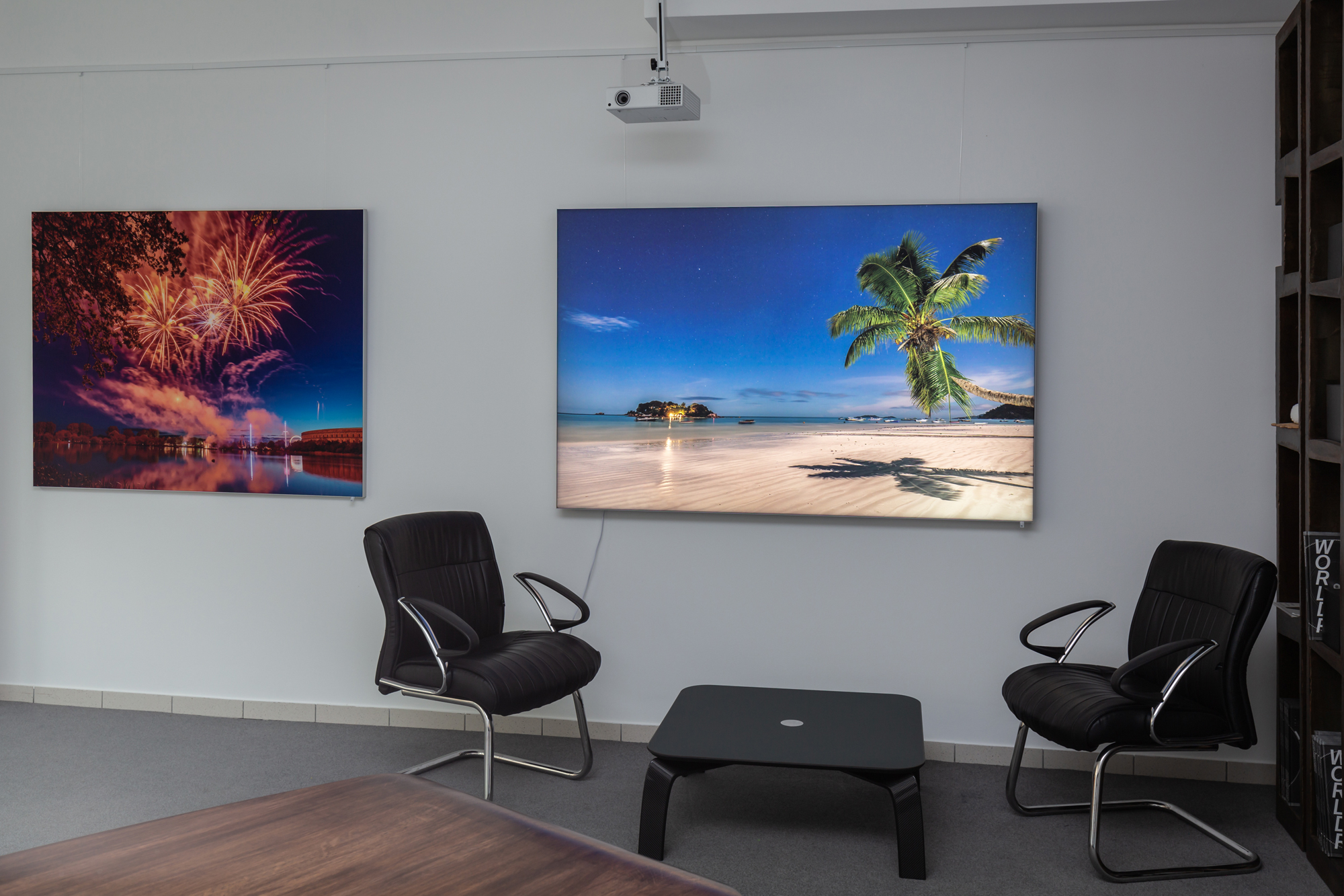 Konferenzraum mit zwei großen Wandbildern: Feuerwerk über Wasser links, Palmenstrand rechts; schwarze Ledersessel.