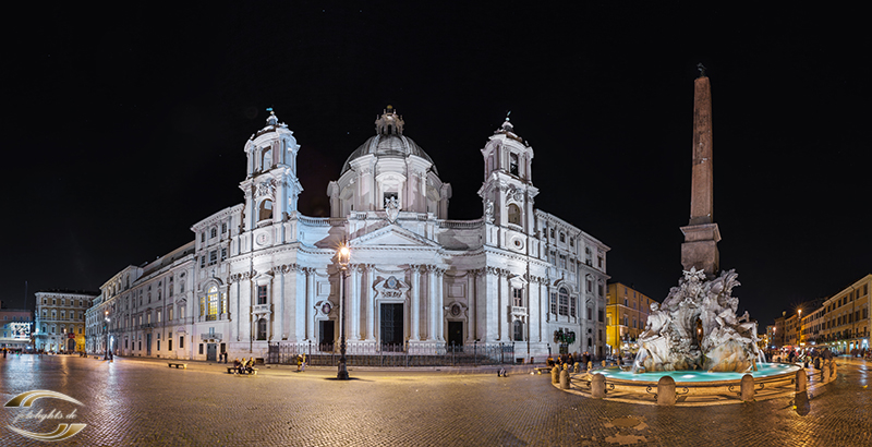 Beleuchtete Barockkirche mit Kuppel und zwei Türmen bei Nacht; rechts Obeliskenbrunnen auf dem Platz.
