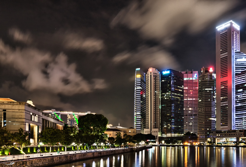 Nächtliche Skyline am Wasser: beleuchtete Hochhäuser, Uferpromenade und Lichtreflexe im Hafen.