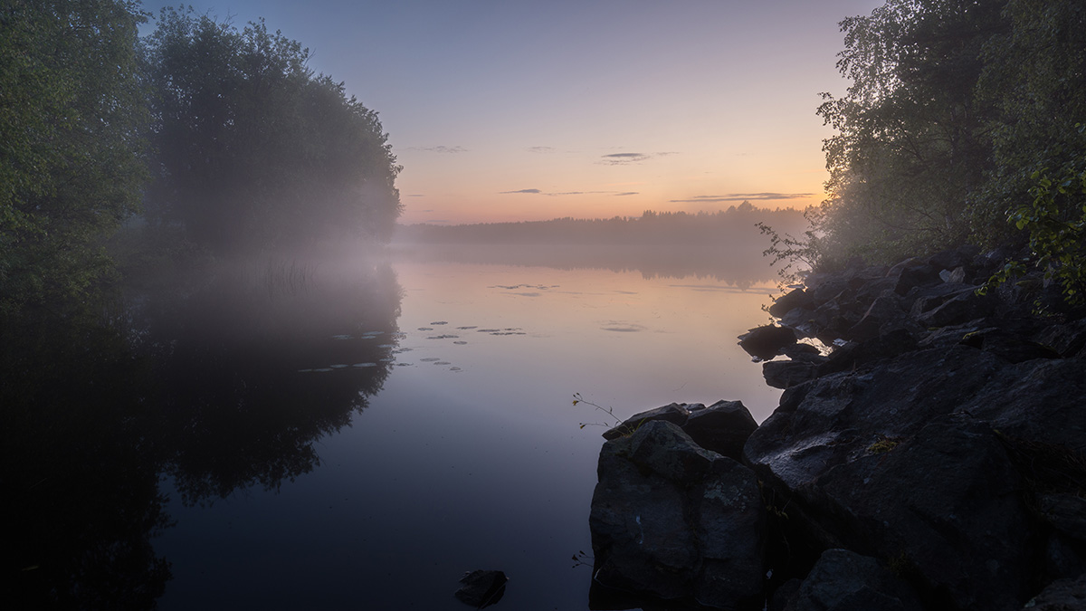 Morgendliche Landschaft mit nebelverhülltem See, umgeben von Bäumen und Felsen, während die Sonne aufgeht und sanfte Farben am Himmel erscheinen.