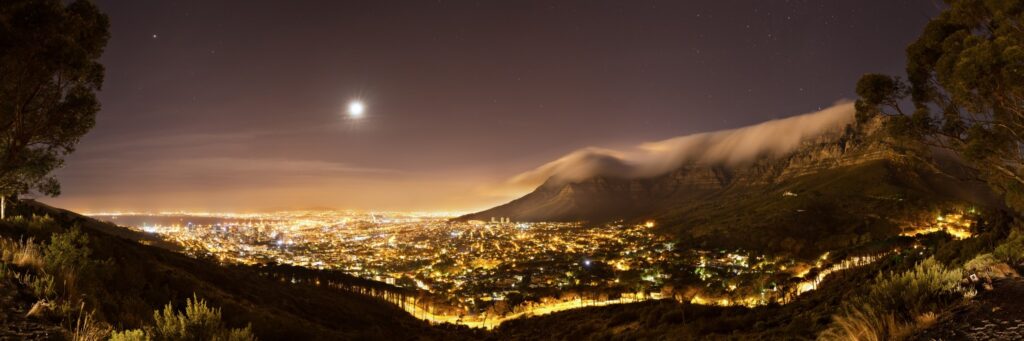 Nächtliche Panoramaaufnahme von Kapstadt: Lichtermeer der Stadt, Mond am Himmel, Wolken über dem Tafelberg.