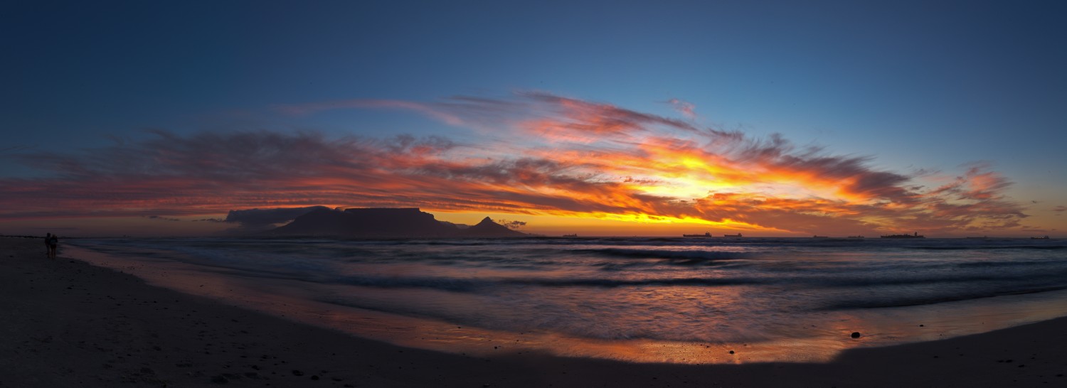 Sonnenuntergang am Strand über dem Meer mit farbigen Wolken und Silhouetten von Menschen am Ufer.
