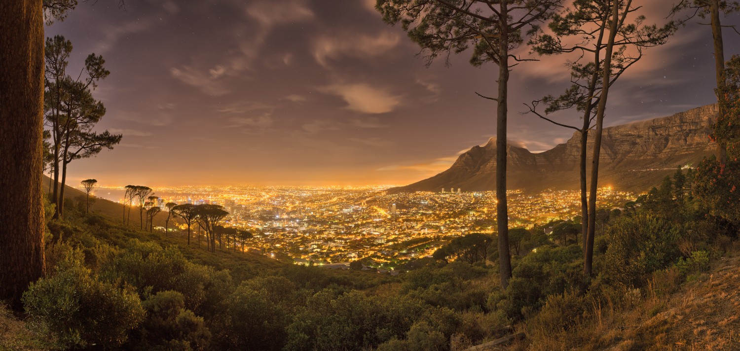 Nachtpanorama einer beleuchteten Stadt von einem Waldhügel mit Bergen im Hintergrund