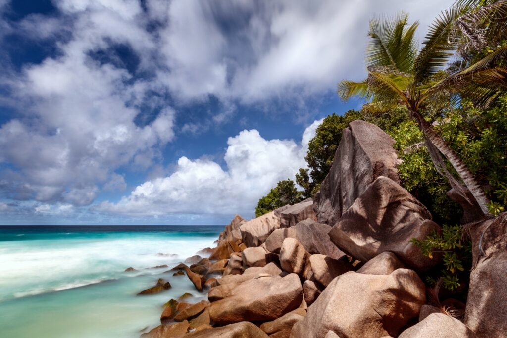 Tropischer Strand mit Felsen, Palmen und türkisblauem Wasser unter blauem Himmel.