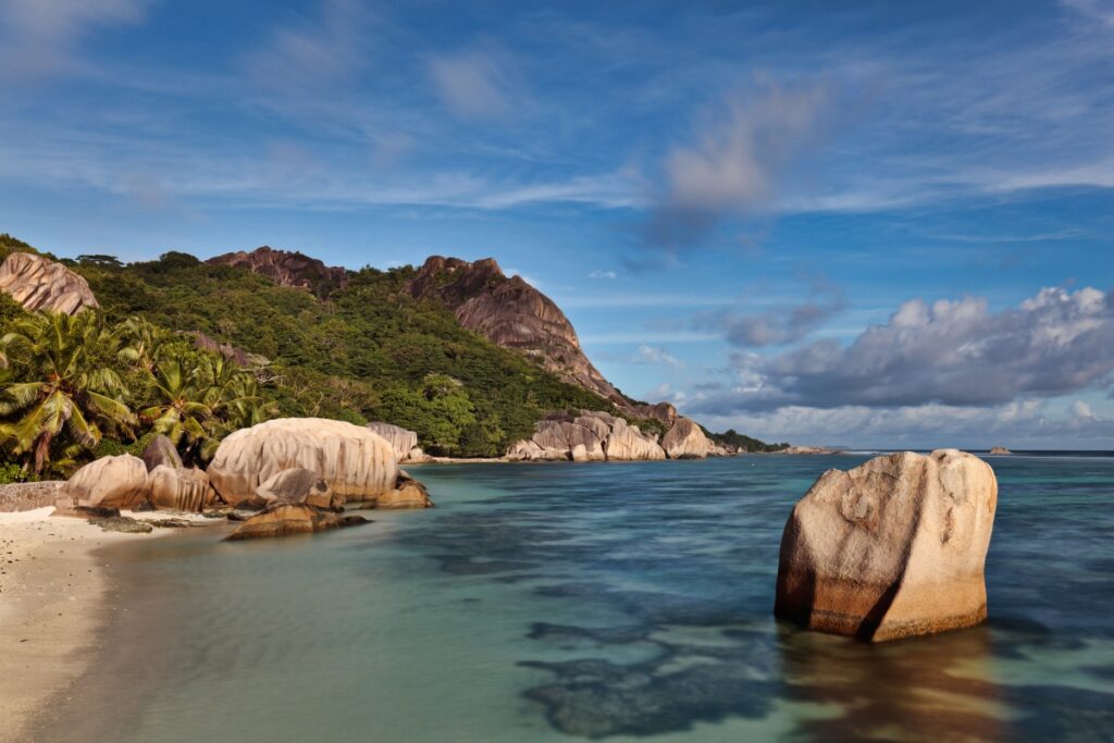 Tropischer Strand mit Palmen, großen Felsen und türkisblauem Wasser vor grüner Küstenlandschaft.