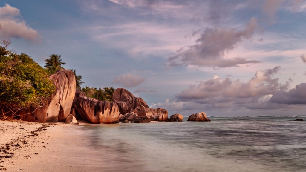 Tropischer Strand mit großen rosa Felsen, Palmen, ruhigem Meer und wolkigem Himmel bei Sonnenuntergang.