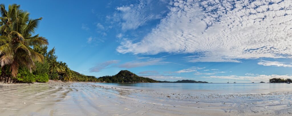 Traumstrand mit Palmen, weißem Sand und türkisblauem Wasser; Inseln im Hintergrund bei blauem Himmel.