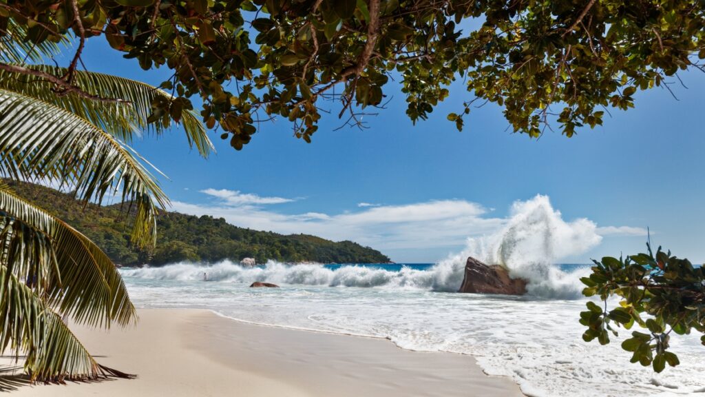 Tropischer Strand mit Palmen, weißem Sand, türkisblauem Wasser und brechenden Wellen gegen Felsen.