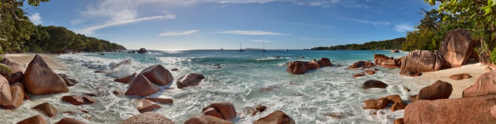 Südseestrand mit großen Felsen, türkisfarbenem Wasser, feinem Sand und Segelbooten am Horizont.
