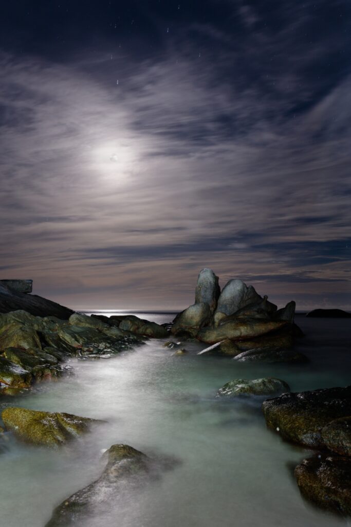 Mondlicht über Felsen an der Küstenlinie, ruhiges Wasser, neblige Wolken – Nacht-Langzeitbelichtung.