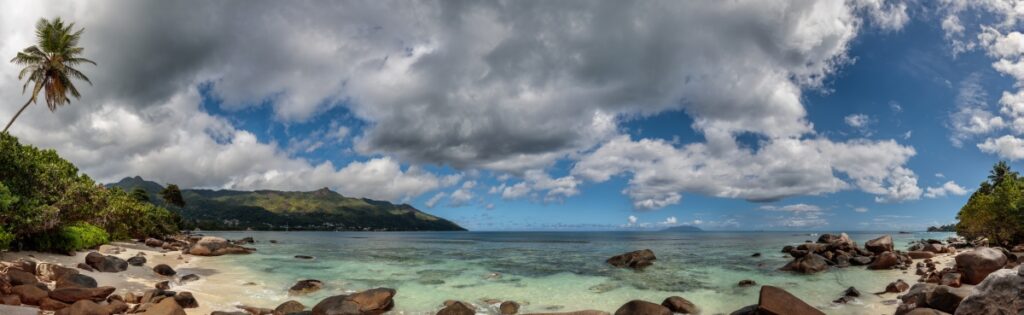 Panorama eines tropischen Strandes mit Palmen, türkisfarbenem Wasser, Sand und Wolkenhimmel.