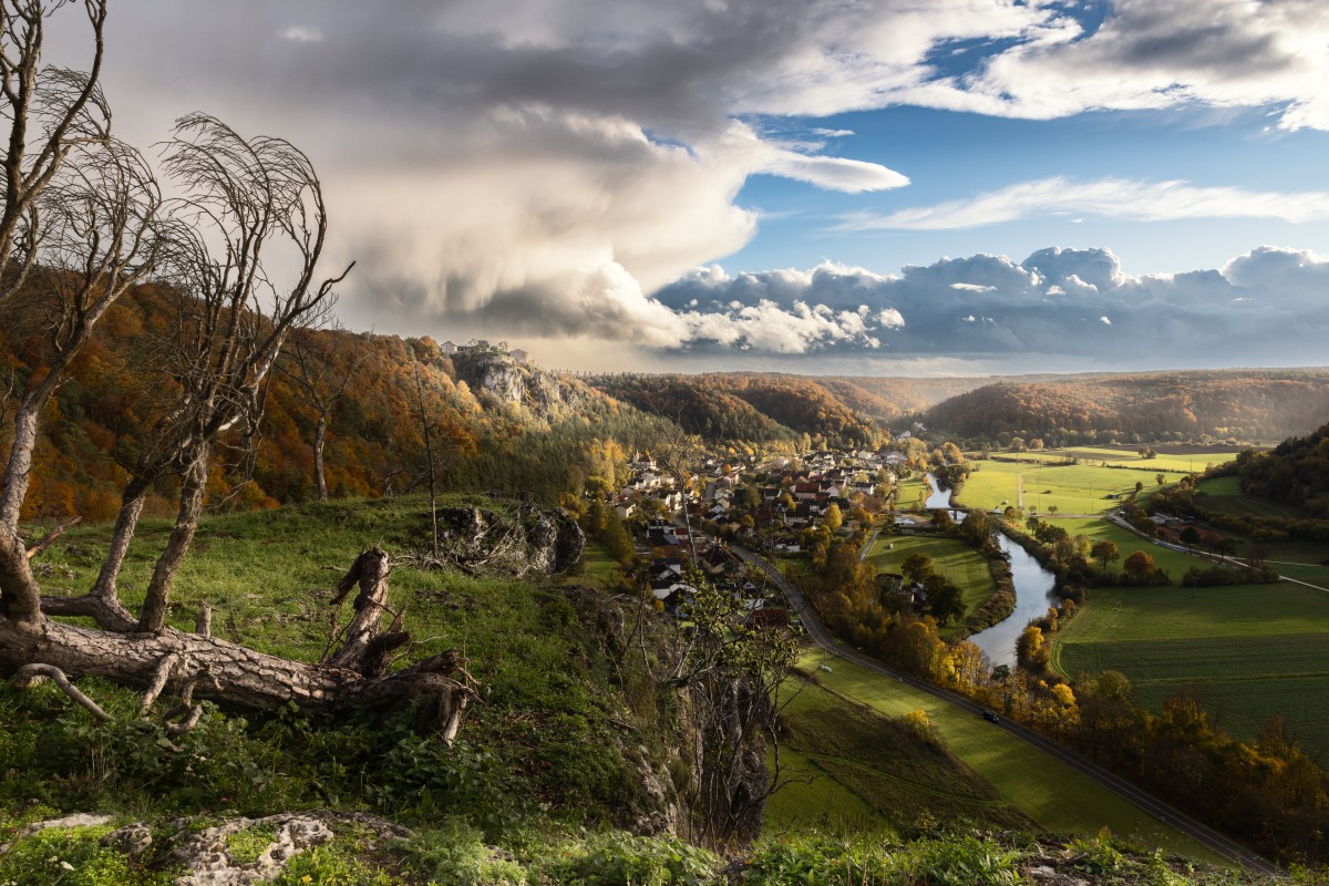 Herbstliche Landschaft: Blick auf Fluss, Dorf und Hügel, umgeben von Wäldern, Felsen und kahlen Bäumen im Vordergrund.