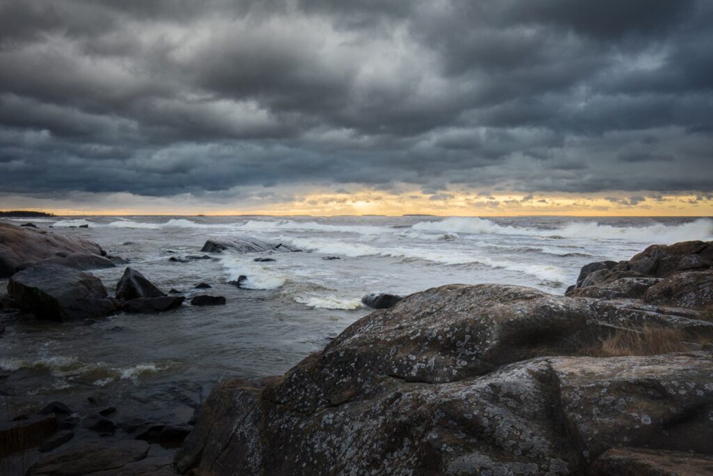 Sturmige Meeresküste mit großen Felsen, tosende Wellen und dunkle Wolken, goldener Horizont am Abend.