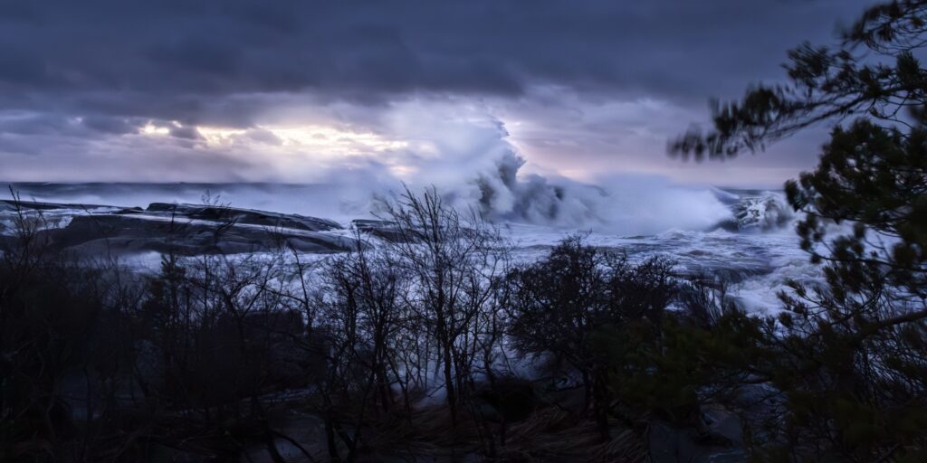 Sturmige Küstenlandschaft: riesige Wellen schlagen gegen Felsen, dunkle Wolken, windgepeitschte Bäume im Vordergrund.