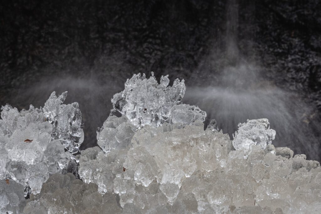 Detailreiche Eiskristalle im Vordergrund; dunkler Hintergrund mit sprühendem Wasserfall dahinter.