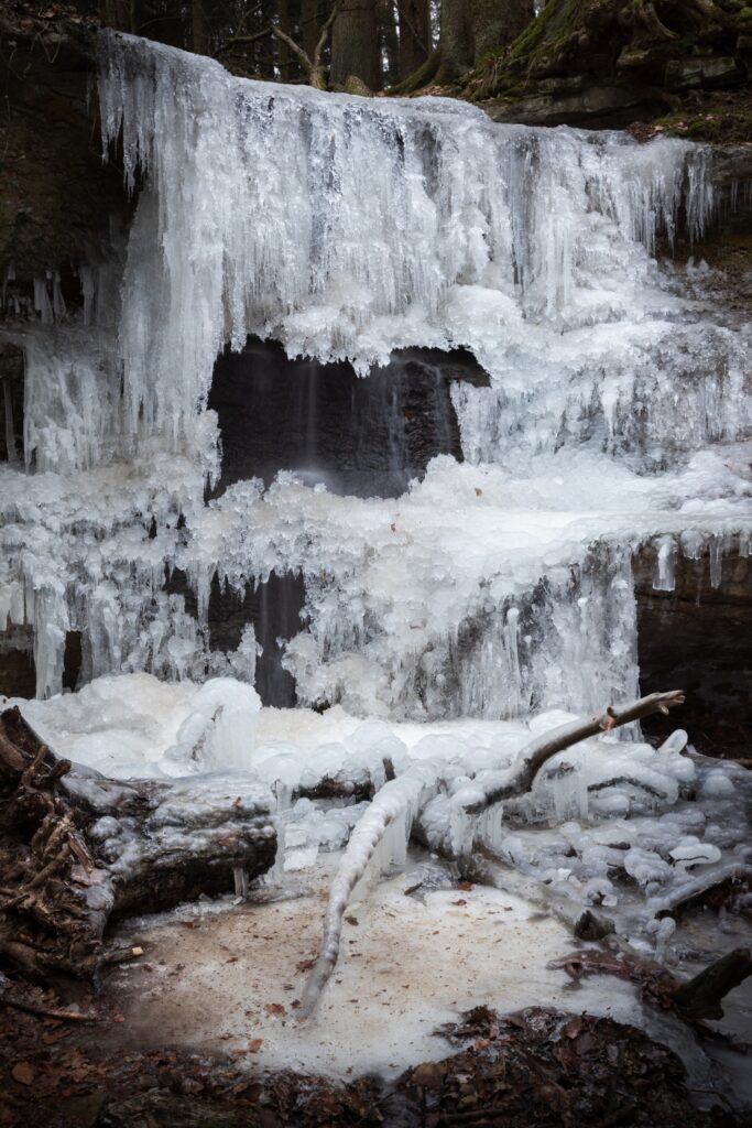 Gefrorener Wasserfall im Wald mit langen Eiszapfen, Eisflächen und umgefallenen Ästen im Winter.