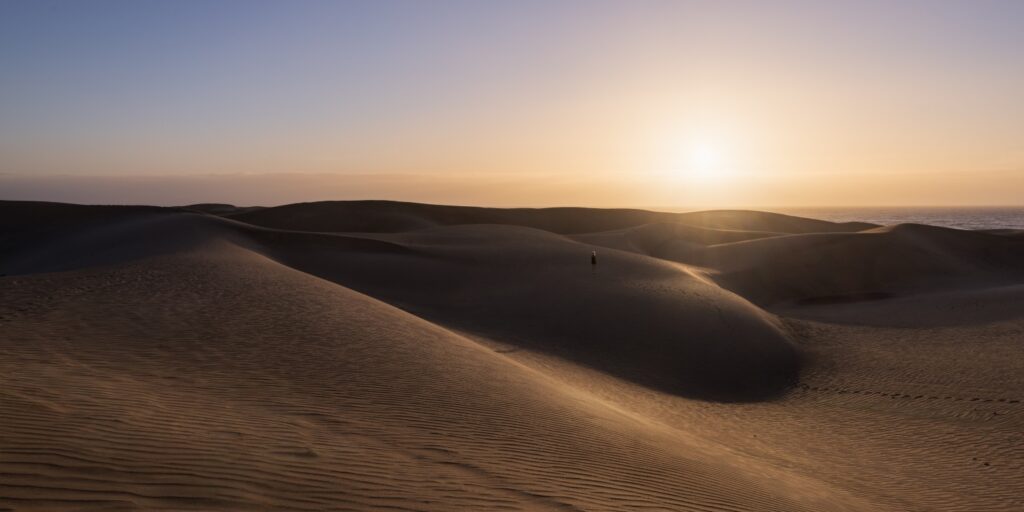 Wüstenlandschaft mit sanften Sanddünen im Sonnenuntergang; einsamer Wanderer am Horizont über dem Meer.