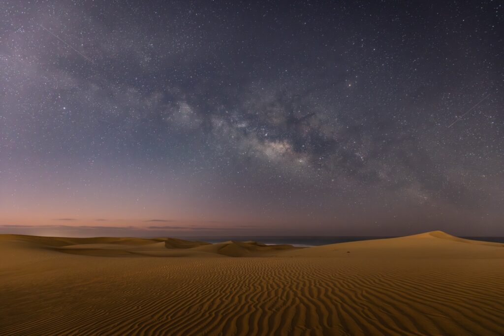 Milchstraße über Wüsten-Dünen bei Nacht, klarer Sternenhimmel und windgeformte Sandmuster.