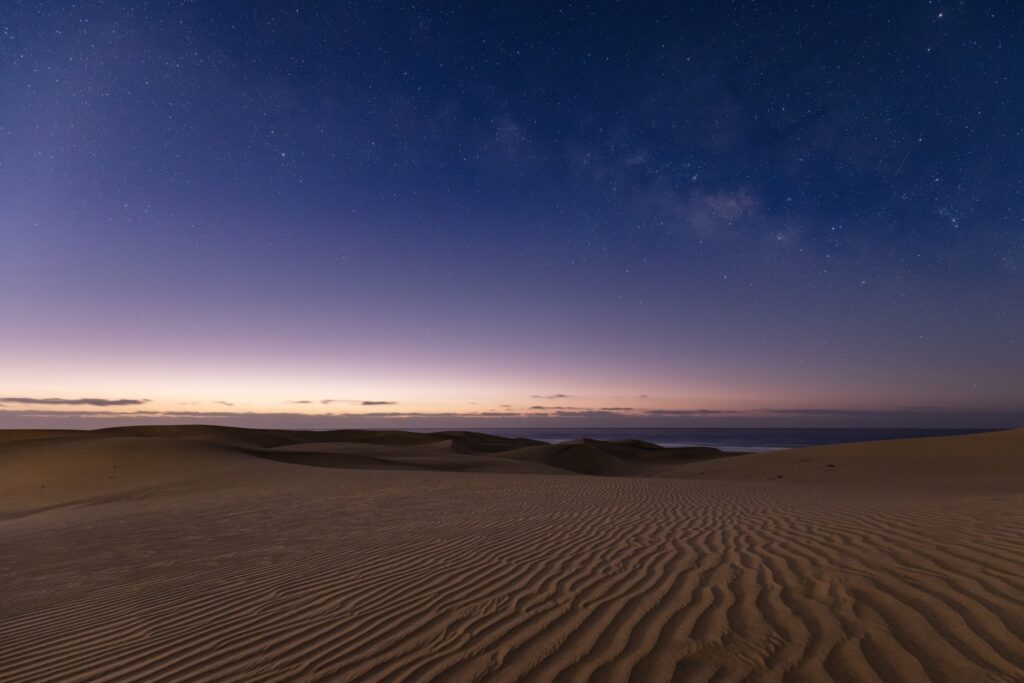 Wüstenlandschaft bei Nacht: Sanddünen mit Rippenmuster, Sternenhimmel und Milchstraße am Horizont.