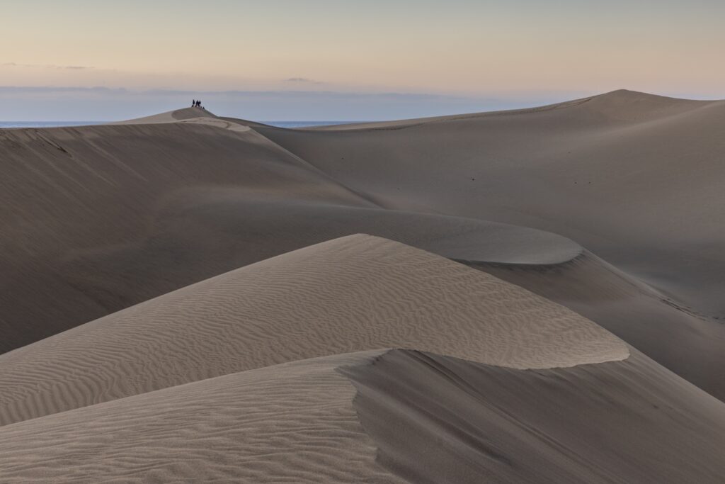 Gruppe von Menschen auf einer Sanddüne am Meer; weite Dünenlandschaft und sanfter Sonnenuntergangshimmel