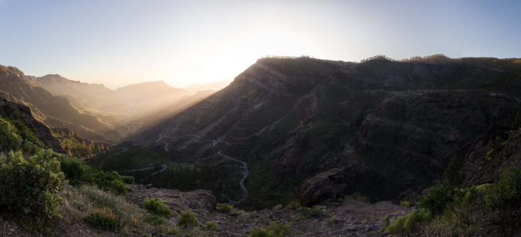 Panoramaaufnahme eines Sonnenaufgangs über einer Schlucht mit kurviger Straße durch grüne Berge.